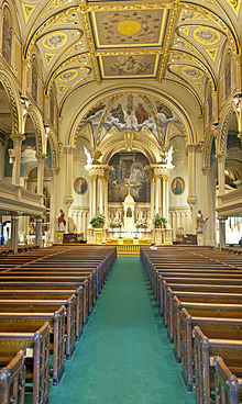 A green-carpeted aisle between two sets of wooden pews with a golden-colored decorated ceiling, arches over the side galleries and an arched, painted nave at the rear