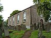 A rectangular grey coloured chapel with large windows and small belfrey sits in a steep churchyard.