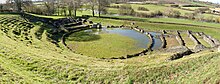 Vue des vestiges d'un théâtre antique adossé à une pente descendant vers un cours d'eau.