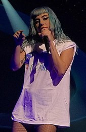 A woman holding a microphone and pointing a finger into audience wearing white shirt