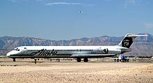 Right side view of an airplane taxiing on the ground towards left side of image. Another plane is behind it, and in the background are mountains and blue sky with a few clouds.