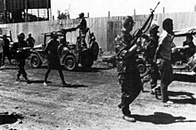 Two men in civilian clothes with their hands on their backs walk surrounded by three armed men in uniform. Military jeeps are seen in a second plane.