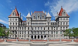 An ornate building, several stories high, of light colored stone. Many arches are visible on its front. On its sides are two large towers with pyramidal red roofs, echoed by similar smaller towers closer to the center with stone tops. In front of the camera, at bottom, is a plaza with a wavy-line pattern.