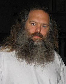 Rubin, sporting a large beard and long hair, and wearing a white tee, stares at the camera without much expression.