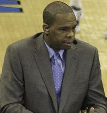 A man seated in a gray suit with a blue tie and dress shirt.