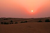 View of dunes at sunset. The setting sun hangs low and dim in a sky graded from fiery orange at top to maroon near the horizon. Terrestrial features are difficult to discern in the crepuscular illumination.