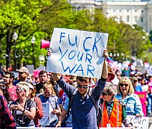 A protester holding a cardboard sign that reads "FUCK YOUR WAR"