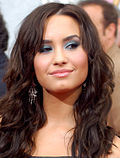 A close-up shot of a teenage girl with long brown hair, looking towards the right and smiling. She is posing at a press event.