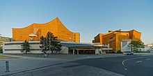 Frontale Farbfotografie von einem Gebäudekomplex im Sonnenuntergang. Das vordere flache Gebäude ist weiß mit einem breiten Eingang. Hinter dem Eingang sind zwei größere Gebäuden in Ockerfarbe mit wellenförmigen Dächern.