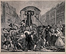 Daniel Defoe stands in a pillory on a high platform. Around him is a thick crowd of people, some of whom are waving their hats and trying to give him flowers. Soldiers around the platform attempt to hold them back.
