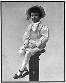 A black-and-white photographed of a neatly-dressed young boy in a hat, sitting on a pedestal.