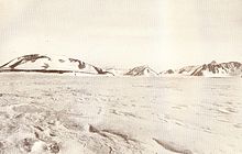 A mountain with a rounded summit and snowy slopes, with smaller hills to its right, rising from flat snow-covered ground