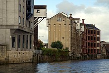 Two riverside mill buildings, each with prominent wooden lucarnes cantilevered beyond their wall above the river, supported by diagonal struts