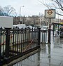 A white sign reading "REGENT'S PARK STATION BAKERLOO LINE" in black letters with people walking behind it all under a light blue sky