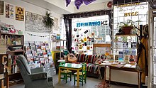 Sunlight shines through the window onto the hangout zone at Spartacus Books, which includes a couch, chair, guitar, colourful magazine rack, posters, and patches.