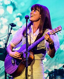 A woman in her late 30s with mid length brown hair and a fringe, wearing a cream blouse that is lightly striped with puffy sleeves and fabric shorts of the same colour and pattern.