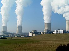 Frontale Farbfotografie von vier Schornsteinen eines Reaktors mit großen Dampfwolken, die die obere Bildhälfte einnehmen. Um die Schornsteine stehen flache Gebäude und zwei Kuppelanlagen. Im Vordergrund sind Felder.