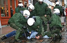 5 police officers in green combat gear with white helmets, partly sitting on a person on the ground.