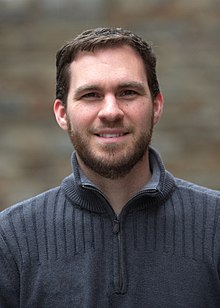 A headshot of Geoff Knorr. He is wearing a sweater and smiling facing the camera with a blurred background.