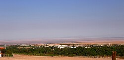 Desert landscape with a broad band of green trees in the foreground, and inmediately behind the trees the town of Pica. Behind the town a large barren plain is seen and farthest parts a series of large but gently.sloping mountains.