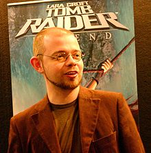 A short-haired man with a beard standing in front of an advertisement poster. The man wears glasses and a light brown jacket over a brown top.