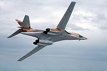 A B-1A flying with its wings swept forward, showing its anti-flash white underside