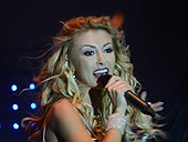 A woman singing into a microphone in front of a blue background.