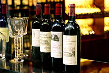 Seitliche Farbfotografie von vier Weinflaschen mit rotem Verschluss und Etiketten, die französischen Text und Gebäudezeichnungen haben. Sie stehen auf einem Glastresen und links neben ihnen sind zwei Weingläser und eine verschwommene Weinflasche. Im verschwommenen Hintergrund sind Regale mit vielen Weinflaschen.