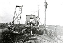 This black and white photograph shows construction workers raising power lines next to the railroad tracks of the Toledo, Port Clinton, Lakeside Railroad tracks in a rural area. The workers are using a railroad car as their vehicle to carry supplies and themselves down the line. It was taken in approximately 1920.