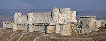 Um castelo de pedra com duas cortinas altas, uma dentro da outra. Elas são enfeitadas e com reentrâncias com torres salientes, tanto as retangulares como as arredondadas. O castelo está em um promontório alto acima da paisagem circundante