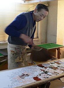 A man hand-dyes a length of white cloth with a floral design, painting in individual flowers in different colours.