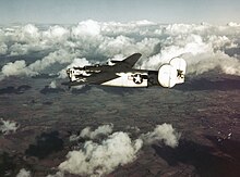 Color photograph of a plane painted white flying above scattered clouds and farmland