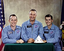 Photographie couleur de l'équipage d'Apollo 1 en combinaison bleue devant une maquette de leur capsule.