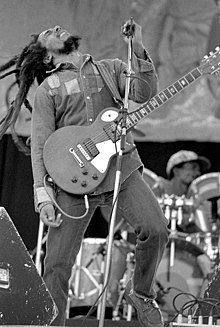 Black and white picture of a man with long dreadlocks playing the guitar on stage.