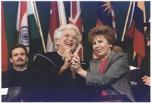 Barbara Bush and Raisa Gorbacheva stand smiling and clasping each other's hands. Bush is wearing a graduation robe.