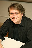 A middle-aged man seated behind a table, facing the camera.