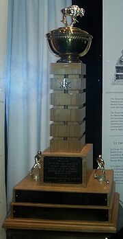 Ornate gold-plated bowl mounted on a wooden plinth atop a two-tier rectangular wooden base adorned with name plates