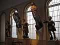An interior view of figures that once graced the bows of wooden sailing ships. High windows flank two female figures that are mounted on the wall, and three male figures stand on pedestals in front of the windows.