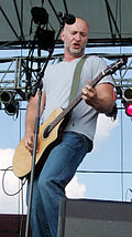 Full length portrait, wearing gray T-shirt and blue jeans, no hair, short gray beard, playing electric guitar.