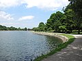 The curving shoreline of a body of water is lined with roughly dressed granite stones. A walkway follows the curve of the shore, with a trees providing shade.