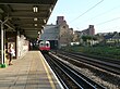 A girl with a pink shirt and blue jeans on a railway platform with a railway track on the right and a white train on it with a red front