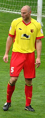 A man wearing a yellow shirt, red shorts and red socks, standing on a grass field. A goalpost and net are visible behind him.