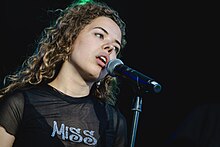 close-up of Nilüfer Yanya wearing a see-thru top that reads "MISS", standing onstage, singing into a microphone