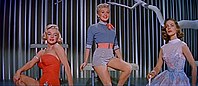 Monroe in How to Marry a Millionaire. She is wearing an orange swimsuit and is seated next to Betty Grable, who is wearing shorts and a shirt, and Lauren Bacall, who is wearing a blue dress.