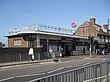 A brown-bricked building with a blue sign reading "ELM PARK STATION" in white letters and people walking in front all under a blue sky