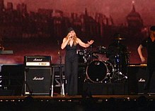 Madonna in a black coat and hat performing in front of a reddish video screen displaying a city skyline
