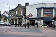 A street with twelve people walking on it and two cars driving on it with three buildings in the background and a grey sky overhead