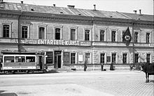 Frontale Schwarzweißfotografie von einer zweigeschossigen Fassade mit dem Banner der Ausstellung „Entartete Kunst“. Links fährt eine Straßenbahn und rechts stehen einige Menschen. Am Eingang befinden sich weitere Tafeln und eine Hakenkreuzflagge hängt am oberen rechten Fenster.