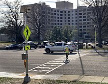 A road with white ladder-style markings and a pole with a yellow, diamond-shaped sign upon which is printed the figure of a walking person.