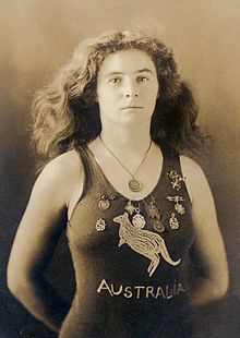 A sepia-toned photograph taken from the waist up, showing a white woman of medium build with shoulder-length wavy hair, wearing a one-piece swimming costume with the word "AUSTRALIA" on the front and a depiction of a kangaroo. She has nine medals pinned to costume around the upper chest area, and is wearing another medal around her neck.